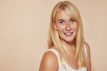 Portrait of a young beautiful woman with freckles smiling and looking at the camera against a beige background