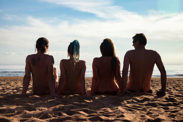 Rear view of silhouettes naked for people men and women sitting on nudist sandy sea beach. From...