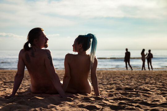 Rear view of silhouettes naked couple man and woman sitting on sandy nudist beach, nature outdoors. Nude young couple relaxing, natural idyllic. Nudism naturism lifestyle concept. Copy ad text space