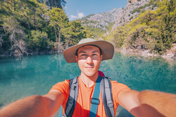 Happy hiker man with trekking backpack taking selfie photo on a trail in mountains with lake in the...