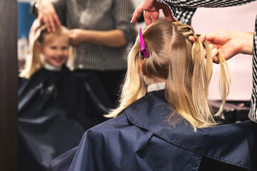 Hairdresser make stylish hairdo for lovely girl in modern barber shop at mirror. Hair salon, barber lady braiding hairdo braids for little blonde kid. Concept of beauty hair care. Copy ad text space
