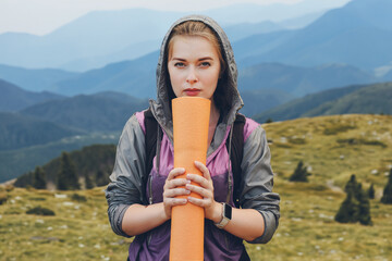 Fototapeta premium A tourist girl with a mat in her hands, dressed in a windbreaker with a hood, looks into the camera against the backdrop of mountain peaks.