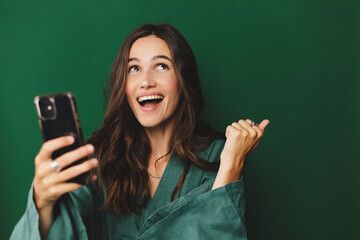Fun young woman hold in hand use mobile cell phone doing winner gesture clenching fists isolated on green background studio. Girl using smartphone apps winning online celebrating discount gift voucher - obrazy, fototapety, plakaty