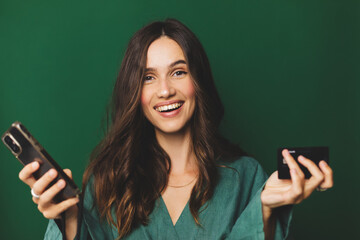 Online shopping. Portrait of smiling brunette curly hair woman paying with plastic credit card on smartphone app, standing with mobile phone and bank card against green background. Happy emotions.