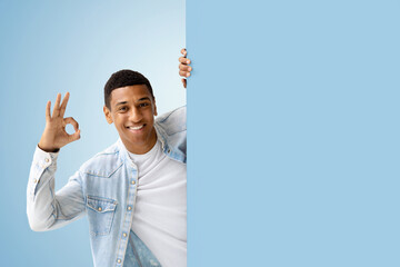 Young African American man peeking out from empty board and shows the OK sign standing on a blue...