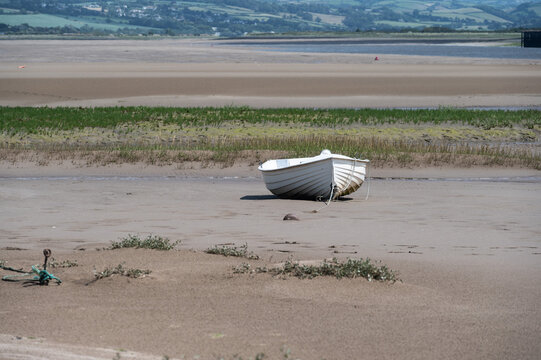 Clinker built, white rowing boat on mud flats at low tide. Sea grass, a sandy beach and green hills provide the background.