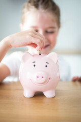 Caucasian child girl putting a coin in a pink piggy bank. Saving money for kids concept. Vertical shot.