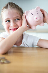 Happy caucasian child girl shaking a smiling pink piggy bank in her hands on a wooden table. Saving money for kids concept. Vertical shot.