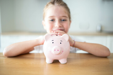 Caucasian child girl holding a smiling pink piggy bank in her hands on a wooden table. Saving money for kids concept.