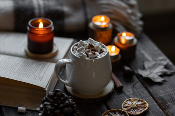 Aromatherapy on a grey fall morning, atmosphere of cosiness and relax. Autumn cozy home composition with hot chocolate with marshmallow and candles. Wooden background, books, close up.