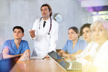 Fototapeta premium Interested adult doctor switching slides with presentation clicker in hand conducting medical consultation with group of colleagues of different ages in medical office..