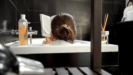 Rear view of elegant woman relaxing in bath with interesting book. Self-care, education, hygiene and spa.
