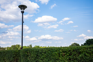 Oświetlenie chodnika w parku w obszarze podmiejskim, na pierwszym planie barierki ochronne, w tle drzewa i błękitne niemalże bezchmurne niebo