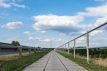 Ścieżka chodnikowa wokół zieleni w okołowiejskich terenach zachodniej Polski w letniej porze przy niemal błękitnym niebem i umiarkowanym zachmurzeniem