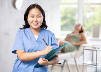 Fototapeta premium Smiling young woman beautician in uniform standing in cosmetology clinic, filling out clipboard with medical records