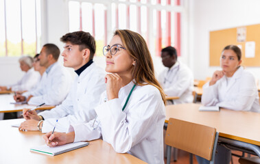Interested young latin american woman in white coat listening to lecture and taking notes in classroom during professional medical training..
