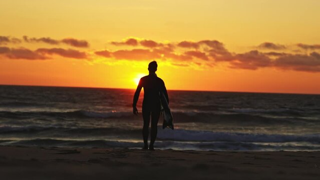 Silhouette of an adult man surfer walking to the ocean at the bright orange sunset