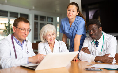 Group of different people in medical uniforms look thoughtfully into laptop standing on table in office