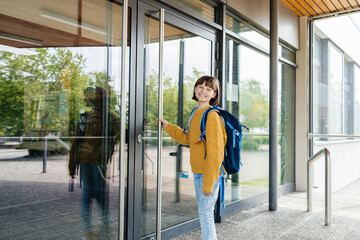 A teenage girl stands in front of the door to the school and opens it. Beginning of lessons and academic year