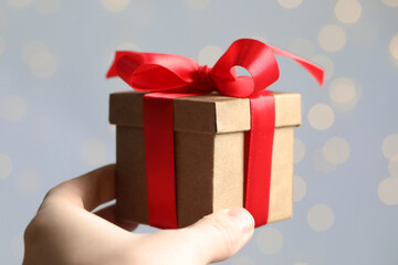 Woman holding gift box with red bow against blurred festive lights, closeup. Bokeh effect