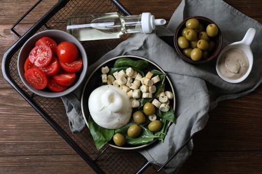 Delicious burrata cheese served with olives, croutons, basil and tomatoes on wooden table, top view