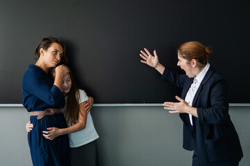 The teacher screams at the schoolgirl and her mother standing at the blackboard. 