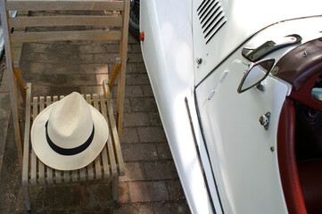 A white vintage car in the sunlight filtering through the trees, a white hat on a wooden chair. 