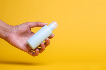 Female hand holding white bottle of body cream on yellow background with copy space for text in right side. Simplicity and commercials concept