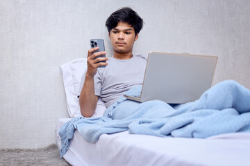 Young busy Asian male sits on bed using laptop to work while reading message from smartphone. Man working on laptop and phone in bed
