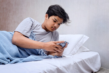 Young Asian man lies in bed under blanket woke up holds smartphone check new messages in morning