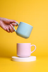 Female hand holding a blue cup and pink cup with white round pedestal on yellow background. 