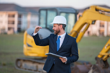 Architect at a construction site. Architect man in helmet and suit at modern home building construction. Architect with a safety vest and suit. Confident architect standing at house background.