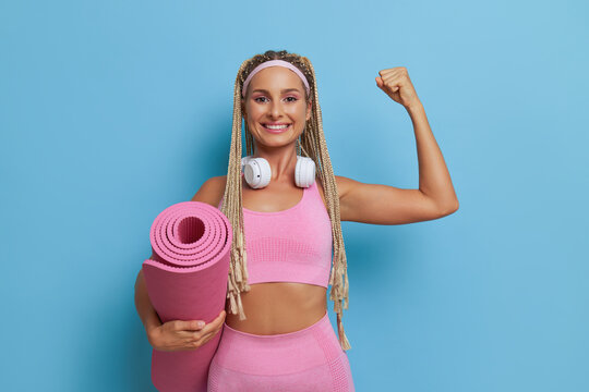 Slender woman in pink top with headphones on her neck stands gainst blue background, holds pink uoga mat and smiles, lifestyle concept, copy space