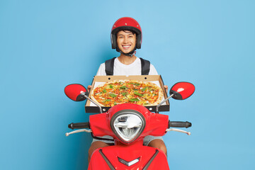 Delivery boy wearing red helmet posing inside studio on his red scooter and holding pizza box in hands, fast delivery concept, copy space