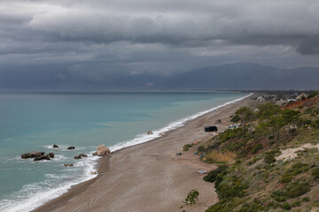Calm seascape on the Mediterranean coast. Cloudy spring weather. Sea water is turquoise. High sloping cliff and a wide sandy beach near the coastline. Mountains and dramatic sky above in the distance