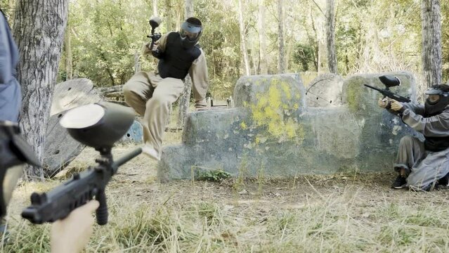 Group of men and women in protective clothes and helmets shooting with paintball markers while hiding behind cover. Teams fighting on battlefield outdoors.