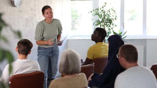 Positive friendly young girl giving lecture to group of women and men of different ages and nationalities during educational class