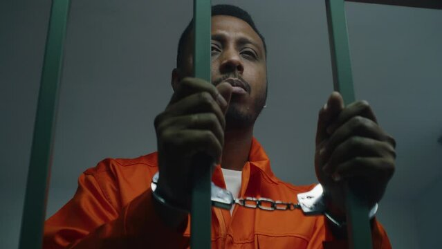 African American man in orange uniform keeps hands in handcuffs on jail cell bars. Depressed murderer serves imprisonment term in prison. Guilty prisoner in correctional facility or detention center.