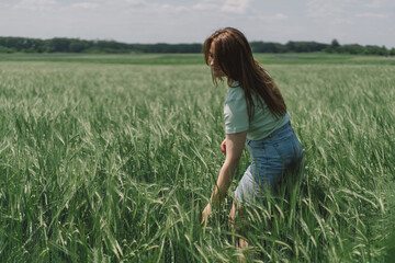 Naklejka na ściany i meble A woman enjoys the fresh air in nature in a green barley field. Summer countryside and gathering flowers. Atmospheric tranquil moment
