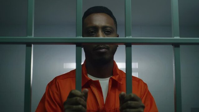 African American prisoner in orange uniform keeps hands in handcuffs on jail cell bars and looks at camera. Guilty criminal serves imprisonment term in prison. Sad murderer in correctional facility.