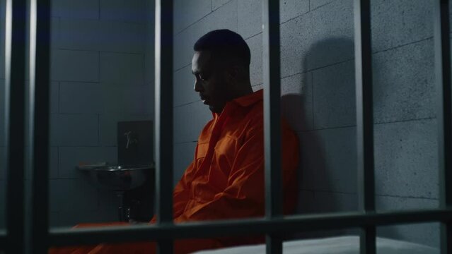 Guilty African American man in orange uniform sits on prison bed and thinks about freedom. Depressed criminal in correctional facility. Sad prisoner serves imprisonment term in jail cell. Dolly shot.