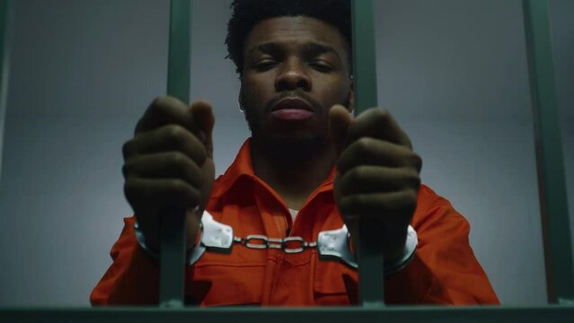 African American man in orange uniform keeps hands in handcuffs on jail cell bars. Depressed murderer serves imprisonment term in prison. Guilty prisoner in correctional facility or detention center.