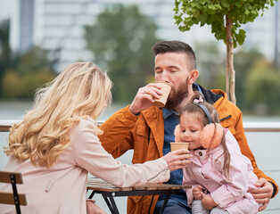 Fototapeta premium Young family man and woman drinking of coffee while spending time with daughter in outdoor cafe. Happy husband and wife sitting at the table with drink cups in new urban district.