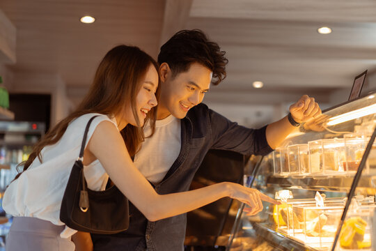 Asian smiling couple looking at cakes through the glass in the bakery and coffee shop. Man and woman pointing and ordering desserts while dating