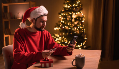 Young man in Santa hat paying online by credit card for Christmas gifts, using smartphone, shopping online, enjoying winter holiday at home office