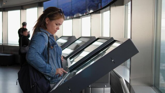 A woman uses a touch-screen terminal in a shopping mall. A woman touches a modern large schedule or information screen in the lobby with large windows.