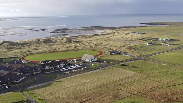 Advancing, wide angle drone shot of the village of Linaclate in Benbecula, featuring the Dark Island Hotel, the local track and football pitch, and the distant beach and ocean. Filmed on the Hebrides.