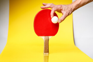 Male hand holding ping pong ball over the red side of a ping pong racket  on a yellow background.