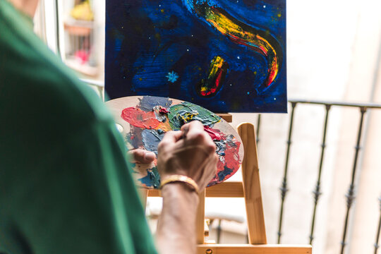 Young man holding artist's palette in front of canvas at home