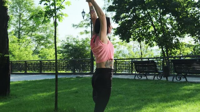 Brown-haired woman raises hands uniting with nature in green park. Lady in pink and black activewear practices balance on yoga class
